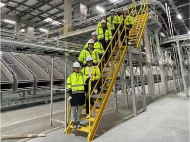 Team of engineers in high-visibility vests posing on yellow stairs overlooking automated warehouse with conveyor systems and storage racks during construction or inspection