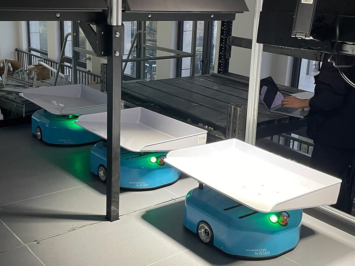 Autonomous mobile robots carrying trays under a conveyor system in a modern warehouse, with a worker using a laptop nearby